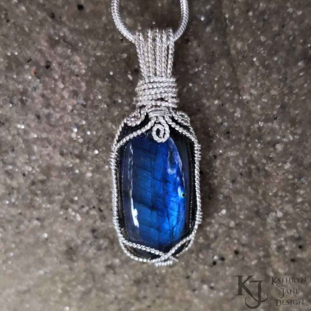 The single twisted silver strand with two anchor points either side of this wee oblong-shaped labradorite provides a simple, open frontal support to display a large amount of the stone's face. The spirals at the top also provide support and symbology for growth.