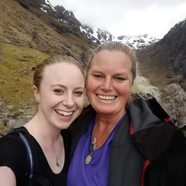 Kathryn relishing being reunited with daughter, Angelique. Coire Gabhail, The Lost Valley, Glen Coe, Scotland. Mother and daughter smiling at the camera. Mother is tanned, having just left New Zealand summer, whereas daughter is pale, having just relished winter in Glasgow, Scotland. Angelique wears amazonite pendant, and Kathryn wears labradorite gemstone crystal and Scottish green stone.