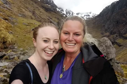 Kathryn relishing being reunited with daughter, Angelique. Coire Gabhail, The Lost Valley, Glen Coe, Scotland. Mother and daughter smiling at the camera. Mother is tanned, having just left New Zealand summer, whereas daughter is pale, having just relished winter in Glasgow, Scotland. Angelique wears amazonite pendant, and Kathryn wears labradorite gemstone crystal and Scottish green stone.