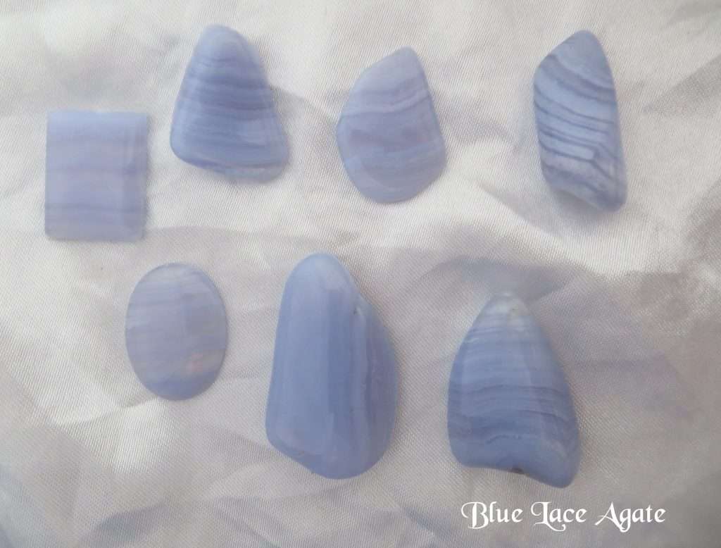 Blue lace agate, polished freeform gemstone crystal shapes. Lilac blue in colour, and opaque, with bands of darker blue and transparency. White satin background.