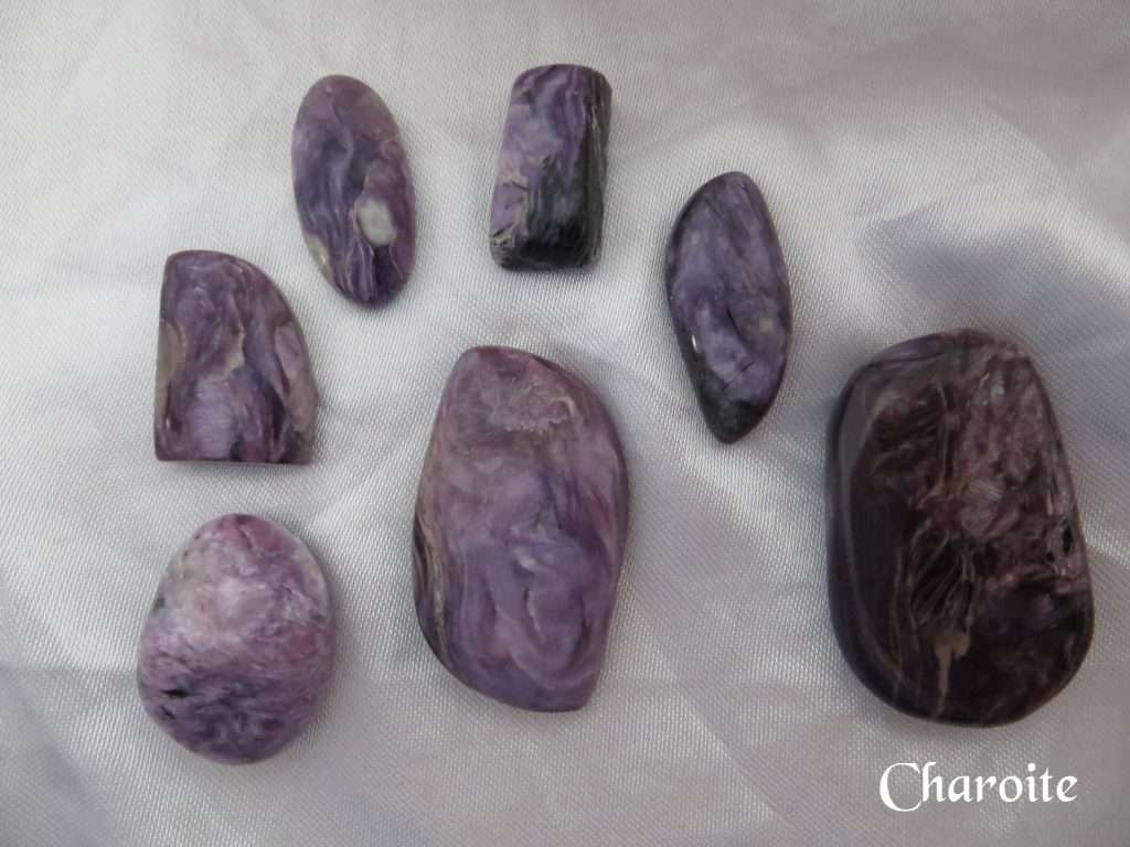 Charoite gemstone crystals. Purple, black, white and lilac colours swirl through various shaped stones. Oval, oblong, freeform, some cabochons, some tumbled. White satin background.