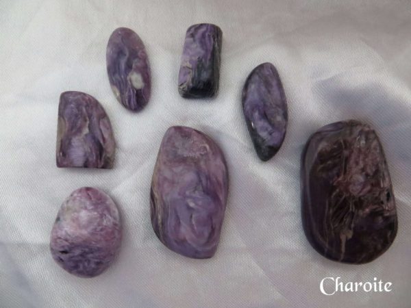 Charoite Charoite gemstone crystals. Purple, black, white and lilac colours swirl through various shaped stones. Oval, oblong, freeform, some cabochons, some tumbled. White satin background.