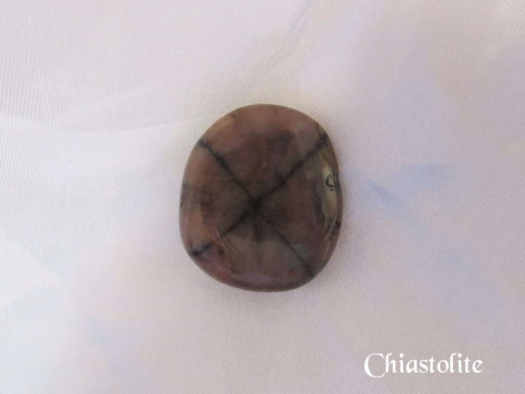 Chiastolite gemstone crystal. Polished stone, slightly round, brown with black cross pattern through entire stone. White satin background.