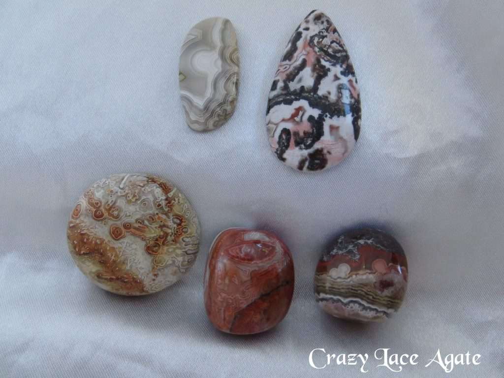 Crazy lace agate gemstone crystals. Cabochons and tumbled stones. Multiple patterns and colours within the stones. Freeform oblong, off-white, looks like partially obscured moon peeking out from behind a section of verticle cloud. Bands of grey, white and brown. A pear shaped cabochon, black, white, brown, salmon-pink random patterns. A circle, orange, brown, yellow, off-white. Two tumbled stone, orange-brown patterns.