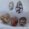 Crazy lace agate Crazy lace agate gemstone crystals. Cabochons and tumbled stones. Multiple patterns and colours within the stones. Freeform oblong, off-white, looks like partially obscured moon peeking out from behind a section of verticle cloud. Bands of grey, white and brown. A pear shaped cabochon, black, white, brown, salmon-pink random patterns. A circle, orange, brown, yellow, off-white. Two tumbled stone, orange-brown patterns.