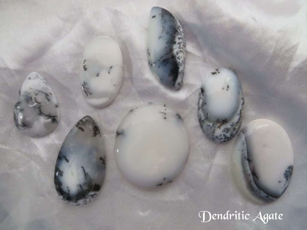 Dendritic agate gemstone crystal. Seven cabochons, shapes including oval, pear and basically round. White stones with black inclusions, and areas of transparency. White satin background.