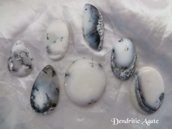 Dendritic agate gemstone crystal. Seven cabochons, shapes including oval, pear and basically round. White stones with black inclusions, and areas of transparency. White satin background.