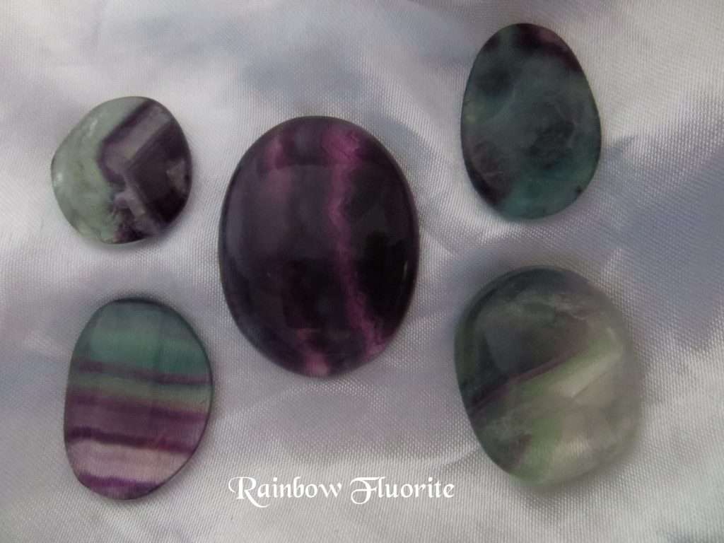 Rainbow fluorite cabochon and polished gemstone crystals. Various shaped gemstones with banded colours of purple, green and transparent on white satin background. All stones have a level of transparency.