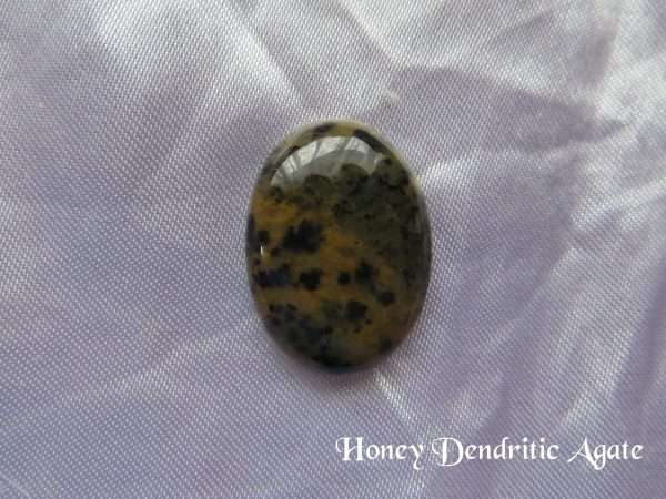 Honey dendritic agate gemstone crystal. Oval cabochon of yellow ochre with black random shapes throughout the stone. White satin background.