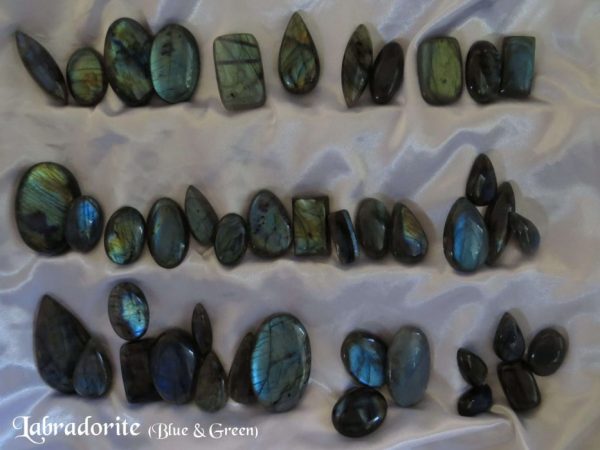 Labradorite cabochons. Large selection which is constantly being replenished. Many labradorite gemstone crystal cabochons, in various shapes and colours of blue and green. White satin background.