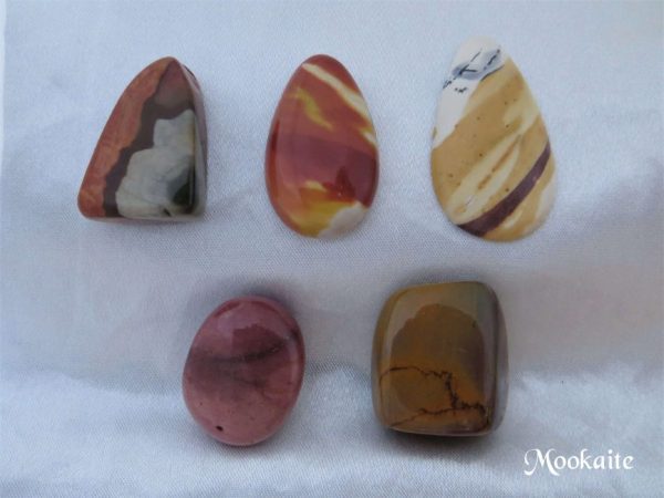 Mookaite, Australia Mookaite gemstone crystals from Australia. Two pear-shaped cabochons and three free-form shaped tumbled and polished stones. Opaque, warm earthy colours of orange, brown, dusky pink, yellow ochre and white. White satin background.