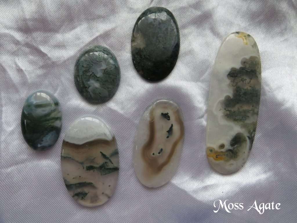 Moss agate gemstone crystals. Six oval cabochons, with various degrees of colour and transparency. Greens, white and wee slivers of brown. They look like moss in water. White satin background.