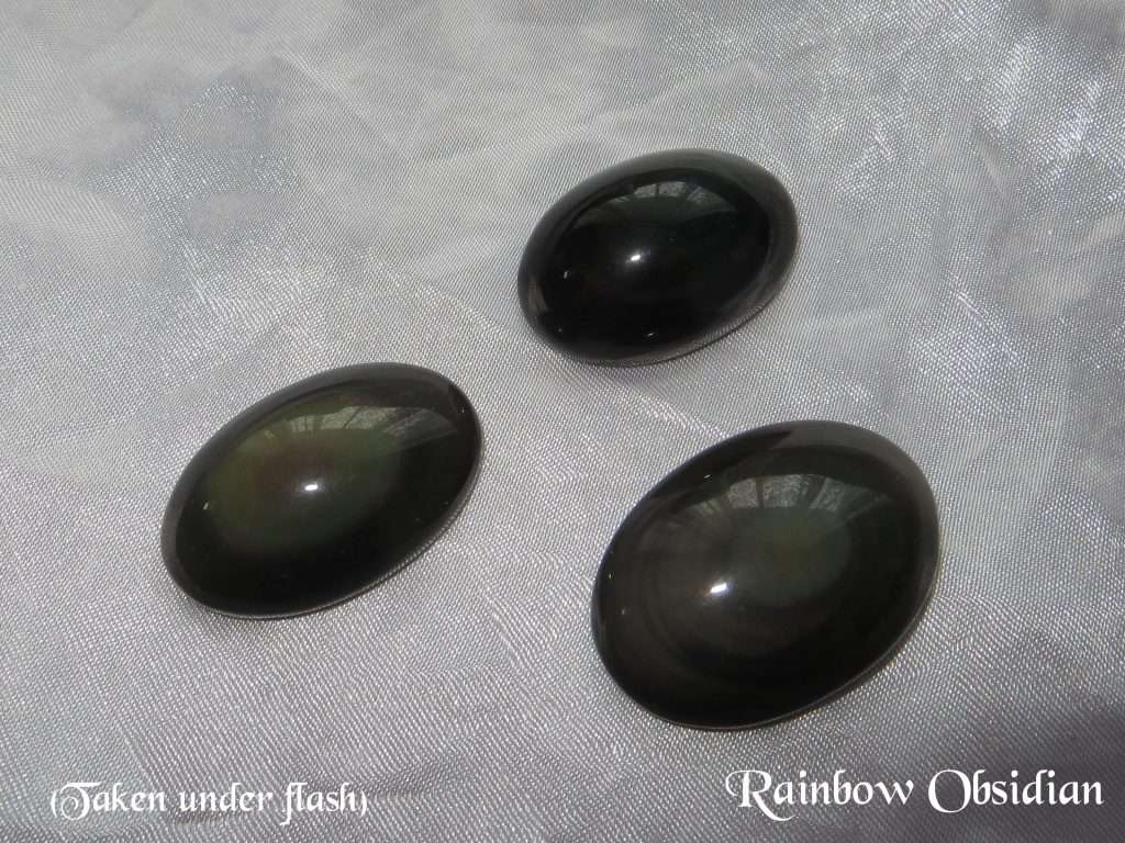 Rainbow obsidian gemstone crystals. Three oval cabochons with rounded tops. Black, opaque, but showing bands of rainbow shimmer near the top. White satin background.
