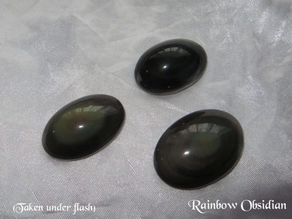 Rainbow obsidian Rainbow obsidian gemstone crystals. Three oval cabochons with rounded tops. Black, opaque, but showing bands of rainbow shimmer near the top. White satin background.