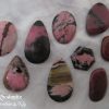 Rhodonite Rhodonite gemstone crystals, from Australia and New Zealand. Nine stones, various shapes and shades of pink with mainly black, some brown, inclusions. Opaque. White satin background.