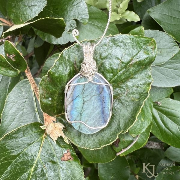 Flashy green, oval labradorite gemstone crystal pendant, lovingly handcrafted into a pendant with sterling silver wire. Reverse side. Green glossy leaves background.