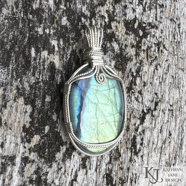 Flashy green, oval labradorite gemstone crystal pendant, lovingly handcrafted into a pendant with sterling silver wire. Twisted wire spiral at top centre. Mossy silvered wood background.