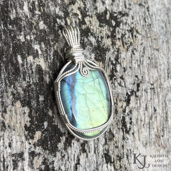 Flashy green, oval labradorite gemstone crystal pendant, lovingly handcrafted into a pendant with sterling silver wire. Twisted wire spiral at top centre. Mossy silvered wood background.