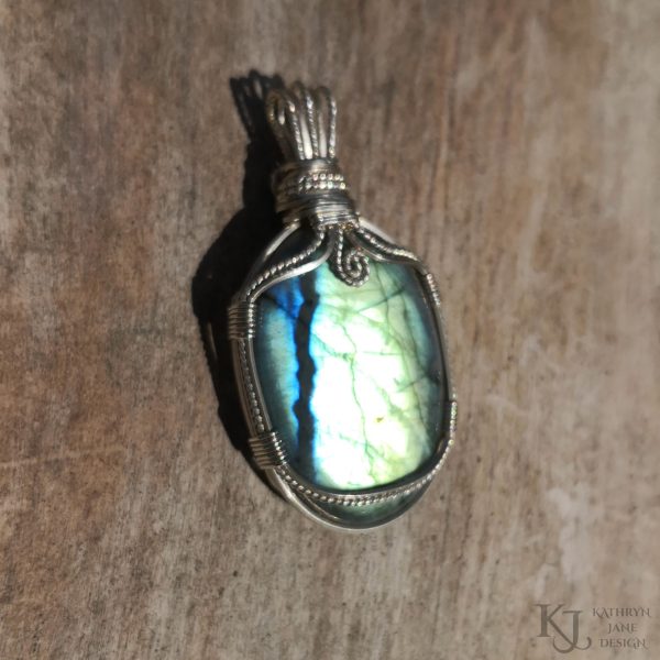 Oval labradorite gemstone crystal pendant. Bright light outdoors, with brilliant light flashing across the face of the stone. Plain wooden background.