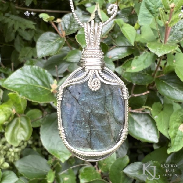 Flashy green, oval labradorite gemstone crystal pendant, lovingly handcrafted into a pendant with sterling silver wire. Twisted wire spiral at top centre. Glossy green leaves background.