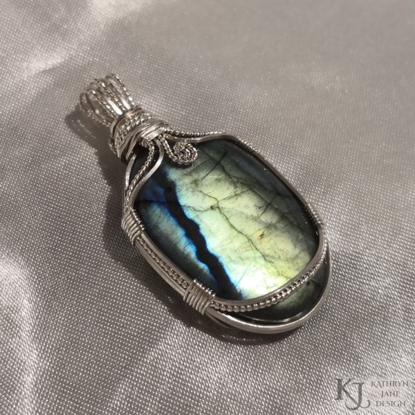 Flashy green, oval labradorite gemstone crystal pendant, lovingly handcrafted into a pendant with sterling silver wire. Twisted wire spiral at top centre. White satin background.