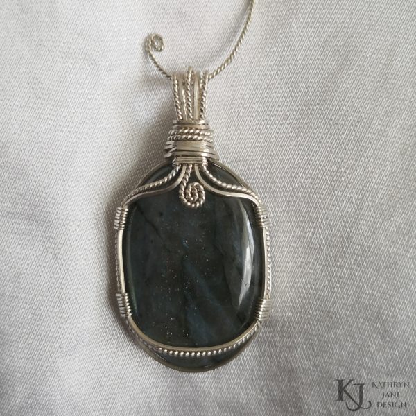 Flashy green, oval labradorite gemstone crystal pendant, lovingly handcrafted into a pendant with sterling silver wire. Glitter sparkles within the stone are showing. Twisted wire spiral at top centre. White satin background.