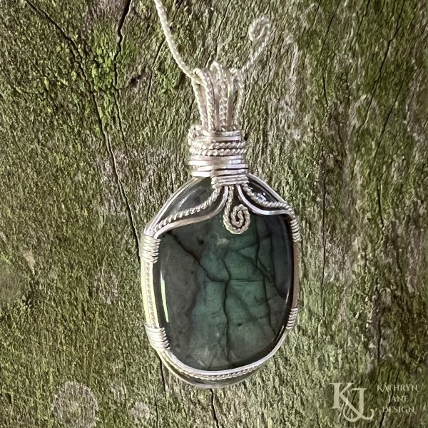 Flashy green, oval labradorite gemstone crystal pendant, lovingly handcrafted into a pendant with sterling silver wire. Twisted wire spiral at top centre. Green mossy wood background.