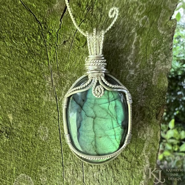 Flashy green, oval labradorite gemstone crystal pendant, lovingly handcrafted into a pendant with sterling silver wire. Twisted wire spiral at top centre. Mossy wood background.
