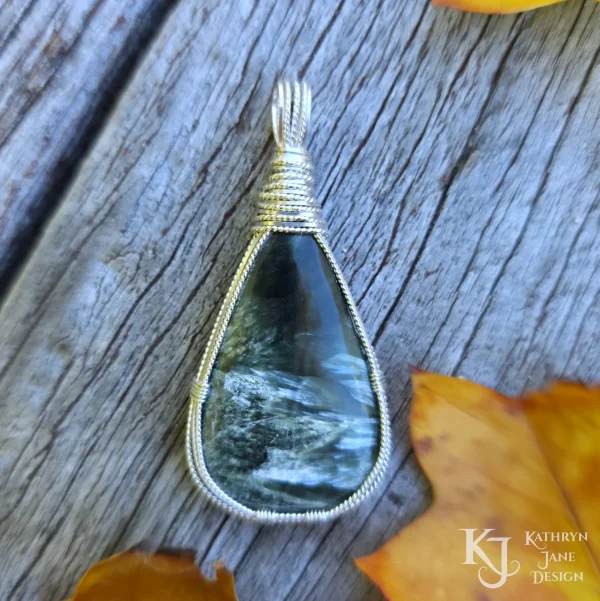 Dark green seraphinite, natural stone, teardrop-shaped pendant, wire-wrapped in twisted sterling silver wire in a simple, outer framework, by Kathryn Jane Design. Wood and golden leaves background.