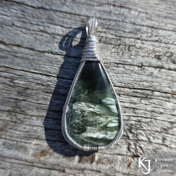 Dark green seraphinite, natural stone, teardrop-shaped pendant, wire-wrapped in twisted sterling silver wire in a simple, outer framework, by Kathryn Jane Design. Wood background.