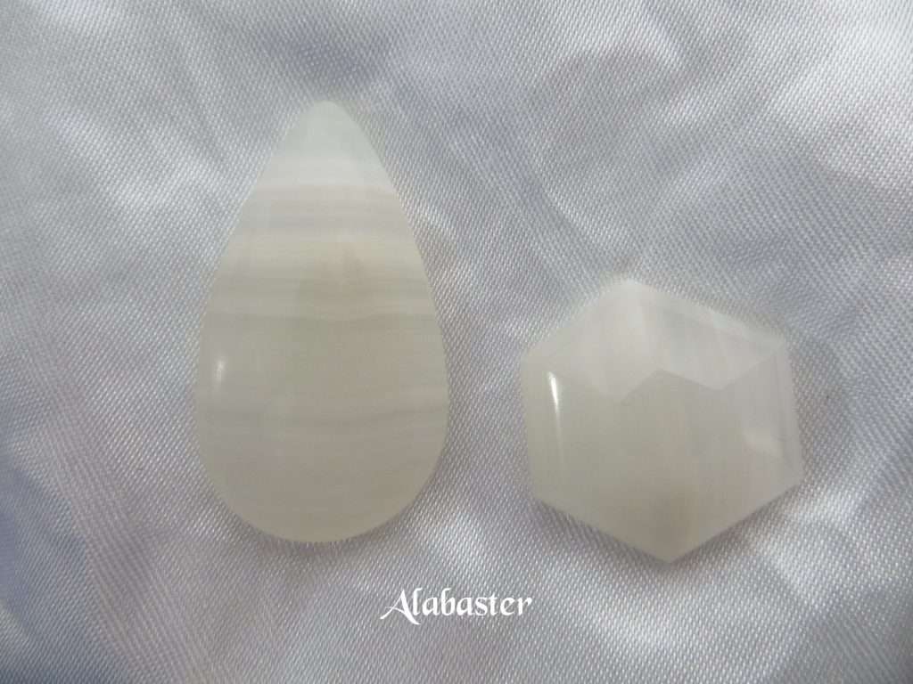 Alabaster gemstone crystal, white cabochons, one a peardrop shape with horizontal bands of various shades of white running through, the other a hexagon. White satin background.