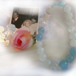 Kathryn created a comforting bracelet of aquamarine and moonstone beads, that felt like a hug from the angels after her parents passed. Tealight candle burns in white praying angel holder. Photo of young girl on mother's knee with father looking on. Pink rose sits on a white display hand, with a bracelet of aquamarine and moonstone gemstone crystal beads draped over the thumb.