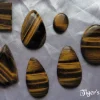 Tiger's eye gemstone crystal cabochons. Shimmers of deep yellow ochre amidst bands of dark brown. Peardrop, oval, oblong and shield shapes. White satin background.