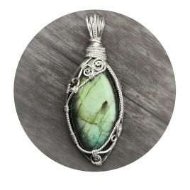 Olive-Green Vesica Piscis-shaped labradorite gemstone crystal pendant. Ornate spirals in sterling silver diagonally across the top and bottom of this beautifully handcrafted, wire-wrapped pendant. Silvered wood background with white circle frame overlay.
