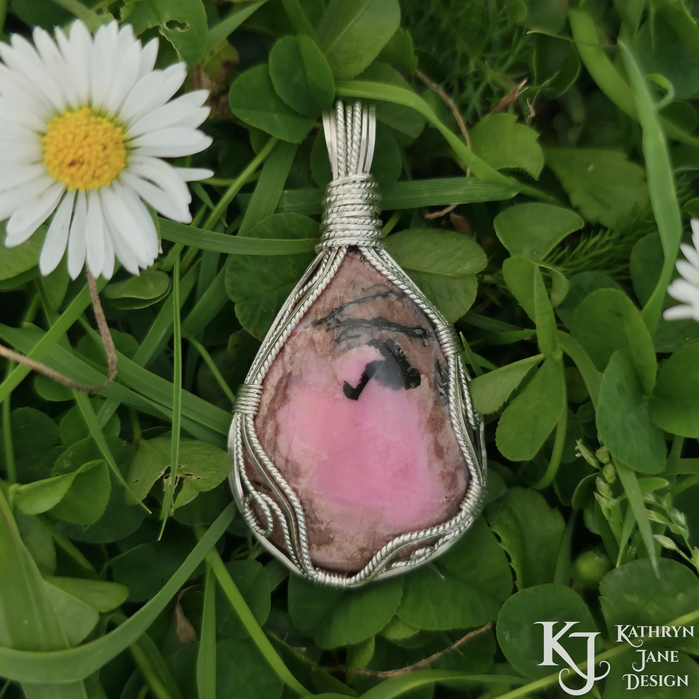 Pink rhodonite pendant, sterling silver wire wrap swirls, on background of green grass, clover and daisy. Kathryn Jane Design "joyous inner child"/
