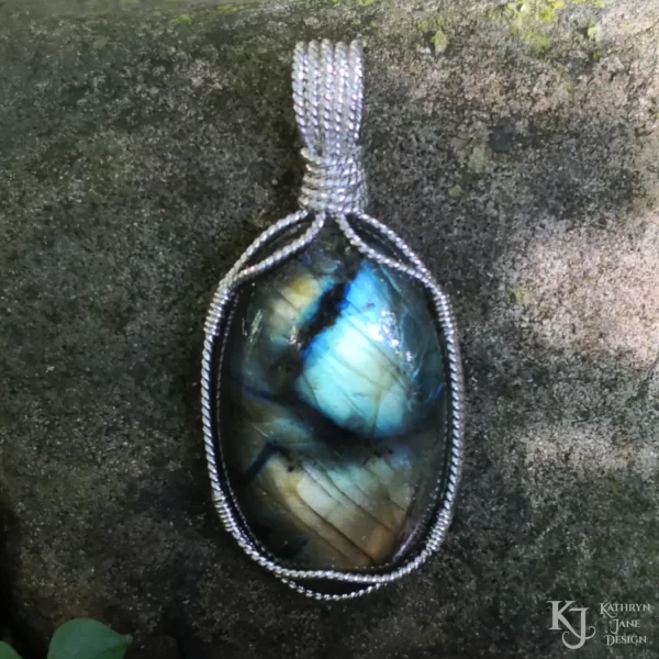 Labradorite gemstone crystal, wire-wrapped in twisted sterling silver, with a natural, moss-covered river stone background. Flashes of blue, green, gold with black branch-like lines running across the face.