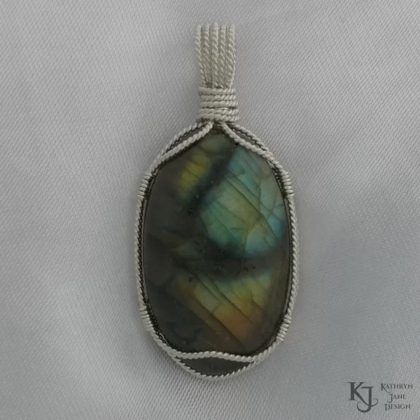 Labradorite gemstone crystal, wire-wrapped in twisted sterling silveron white satin background. Flashes of blue, green, gold with black branch-like lines running across the face.