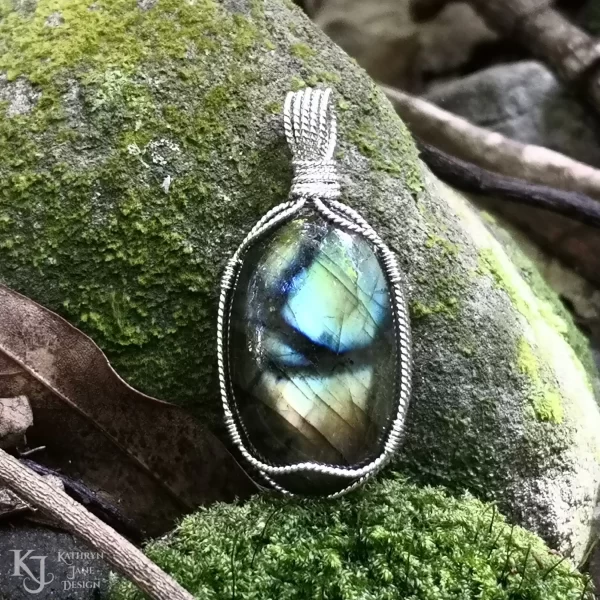 Labradorite gemstone crystal, wire-wrapped in twisted sterling silver, sitting on a bed of moss, with a natural, moss-covered river stone background. Flashes of blue, green, gold with black branch-like lines running across the face. Leaf litter and twigs in foreground and background.