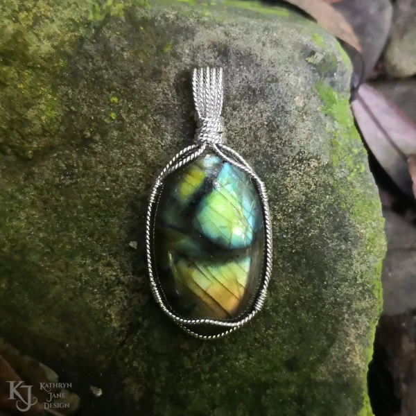 Labradorite gemstone crystal, wire-wrapped in twisted sterling silver, on a natural, moss-covered river stone. Flashes of blue, green, gold with black branch-like lines running across the face.