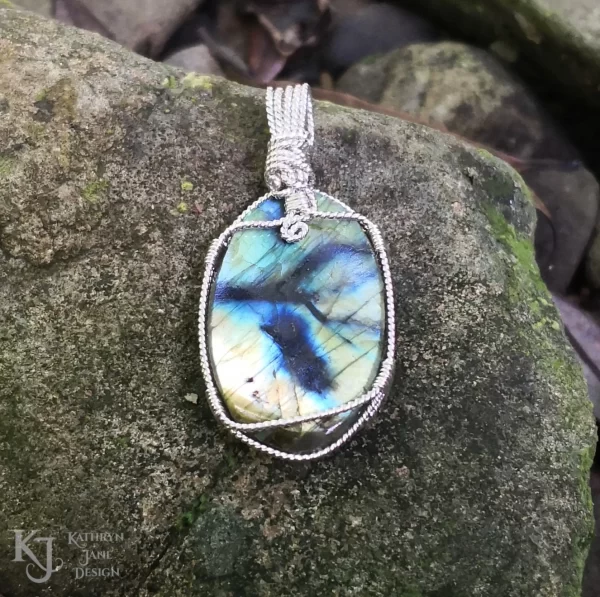 Labradorite gemstone crystal, wire-wrapped in twisted sterling silver, with a natural, moss-covered river stone background. Flashes of blue, green, gold with black branch-like lines running across the reverse side. In Kathryn's imagination, a guardian angel was seen in the inky blue and black lines.