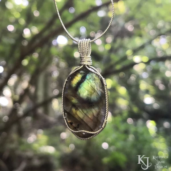 Labradorite gemstone crystal, wire-wrapped in twisted sterling silver, with dappled light on leaves and criss-crossing branches background. Flashes of blue, green, gold with black branch-like lines running across the face.