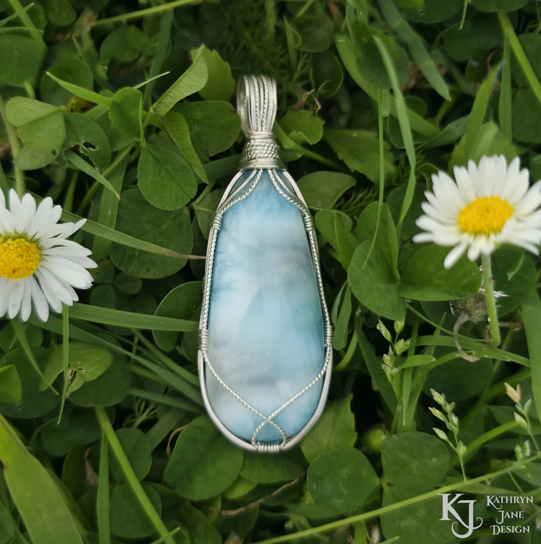 Turquoise-blue larimar pendant, with square and twisted sterling silver wire support and bail by Kathryn Jane Design. Green clover background with two white daisies with yellow centres.