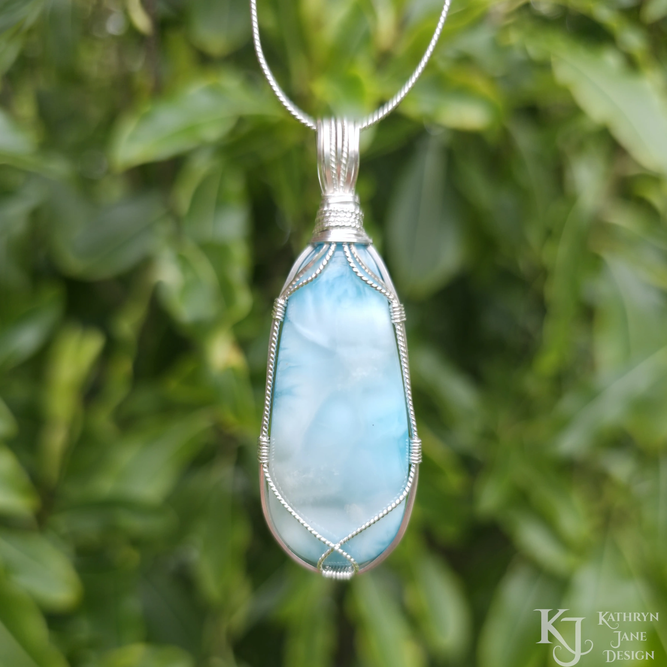 Turquoise-blue larimar pendant, with square and twisted sterling silver wire support and bail by Kathryn Jane Design. Green leaves in background