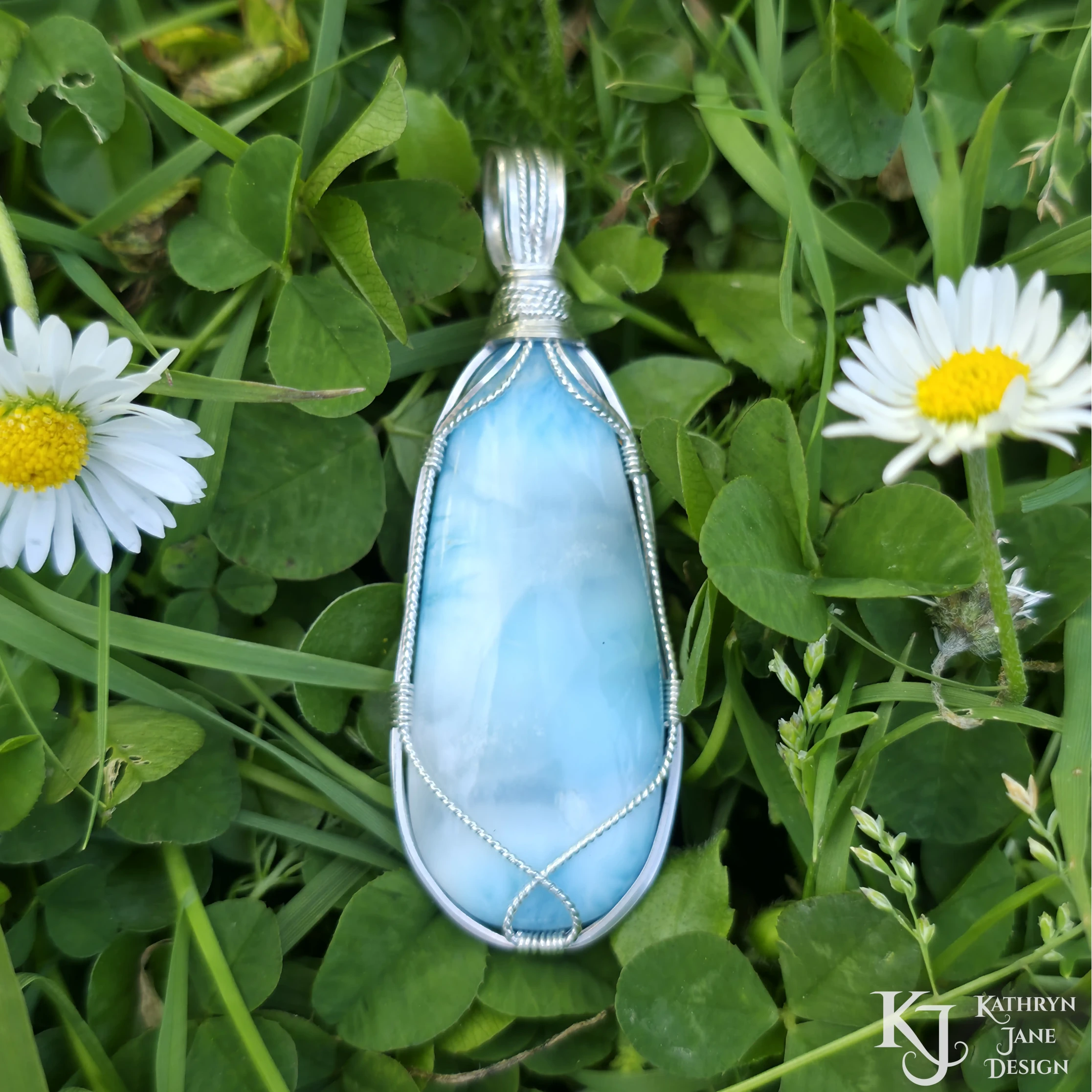 Turquoise-blue larimar pendant, with square and twisted sterling silver wire support and bail by Kathryn Jane Design. Green clover background with two white daisies with yellow centres.
