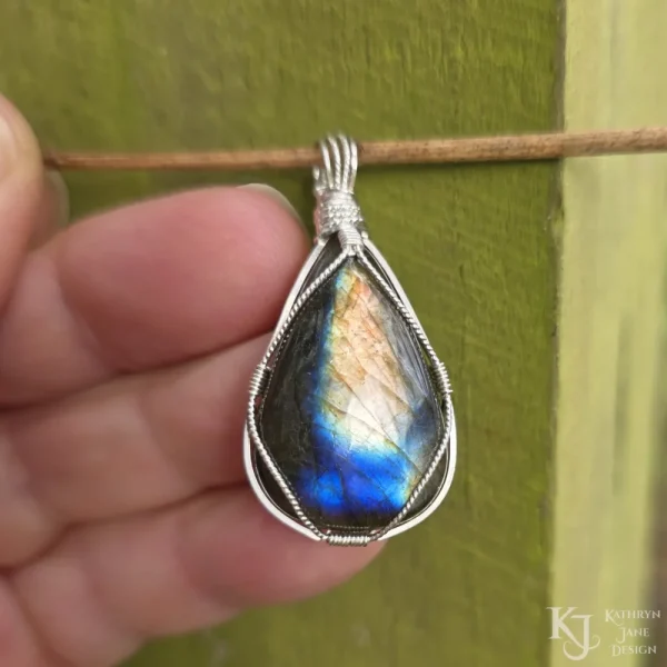 In bright, natural light outdoors Triangular-shaped natural labradorite pendant in a simple sterling silver framework in hand in bright outdoor ight. Labradorescence showing brilliant gold and electric blue