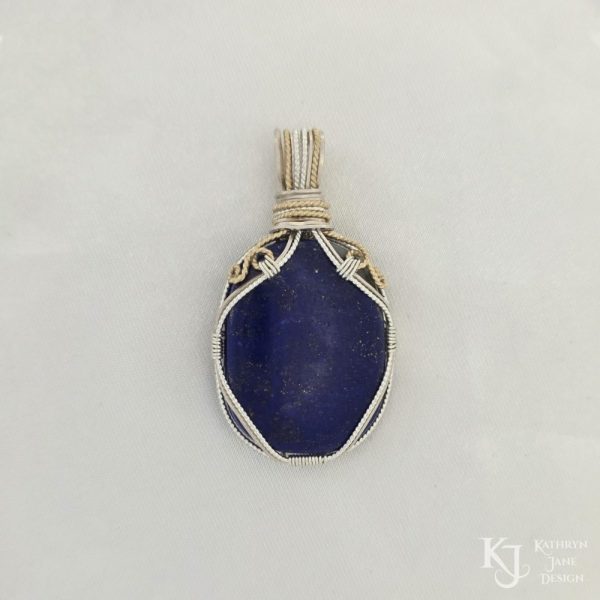 Photo taken in filter cone, eliminating the appearance of reflections off the highly polished surface of the stone Oblong-shaped royal blue lapis lazuli pendant with sterling silver and 14k gold-filled wire spirals at the top, and in the bail. White satin background with Kathryn Jane Design logo in the bottom right