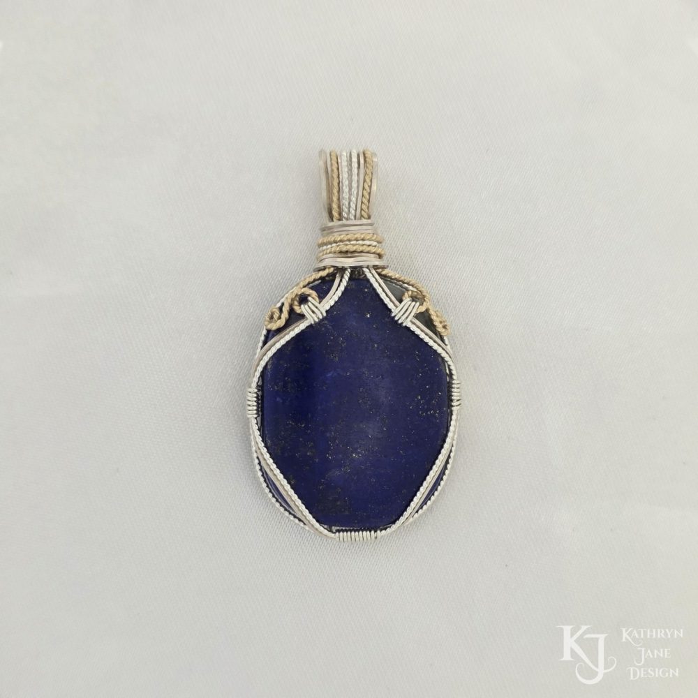 Oblong-shaped royal blue lapis lazuli pendant with sterling silver and 14k gold-filled wire spirals at the top, and in the bail. White satin background with Kathryn Jane Design logo in the bottom right