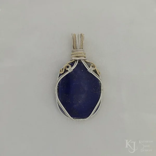 Photo taken in filter cone, eliminating the appearance of reflections off the highly polished surface of the stone Oblong-shaped royal blue lapis lazuli pendant with sterling silver and 14k gold-filled wire spirals at the top, and in the bail. White satin background with Kathryn Jane Design logo in the bottom right