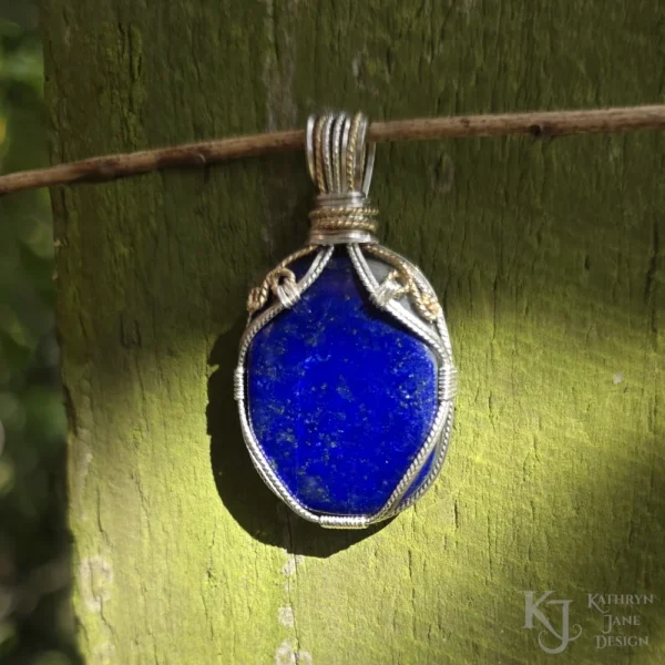 The direct sunlight shows the richness of the blue colouration. See the white calcite shoulder underneath the gold-filled spiral? Oblong-shaped royal blue lapis lazuli pendant with sterling silver and 14k gold-filled wire spirals at the top, and in the bail. Rich royal blue colour visibile in direct sunlight, as well as white calcite shoulder, with Kathryn Jane Design logo in the bottom right