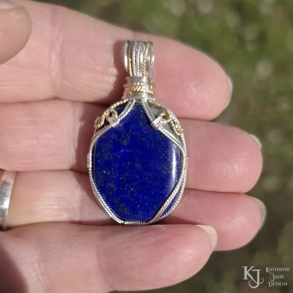 In direct sunlight outdoors Oblong-shaped royal blue lapis lazuli pendant with sterling silver and 14k gold-filled wire spirals at the top, and in the bail. Held in hand in direct sunlight